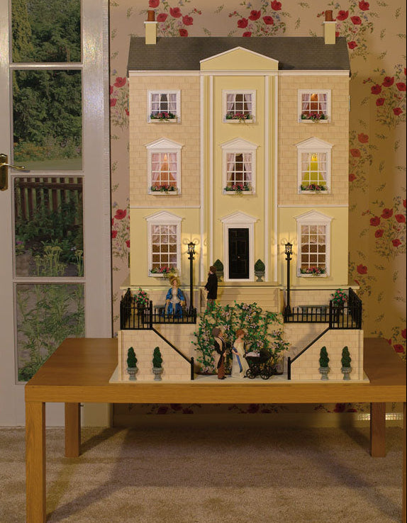 A miniature dollhouse-style house with a cream facade, gray roof, and black railing, displayed on a wooden table in a room with floral wallpaper and French doors.