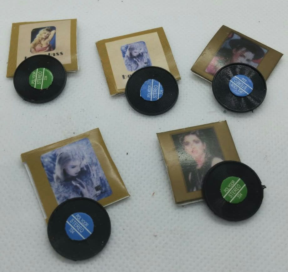 five miniature collectible items arranged on a white textured surface, featuring small portrait photo cards, black vinyl record-shaped pieces with colored centers, and gold-toned rectangular pieces.
