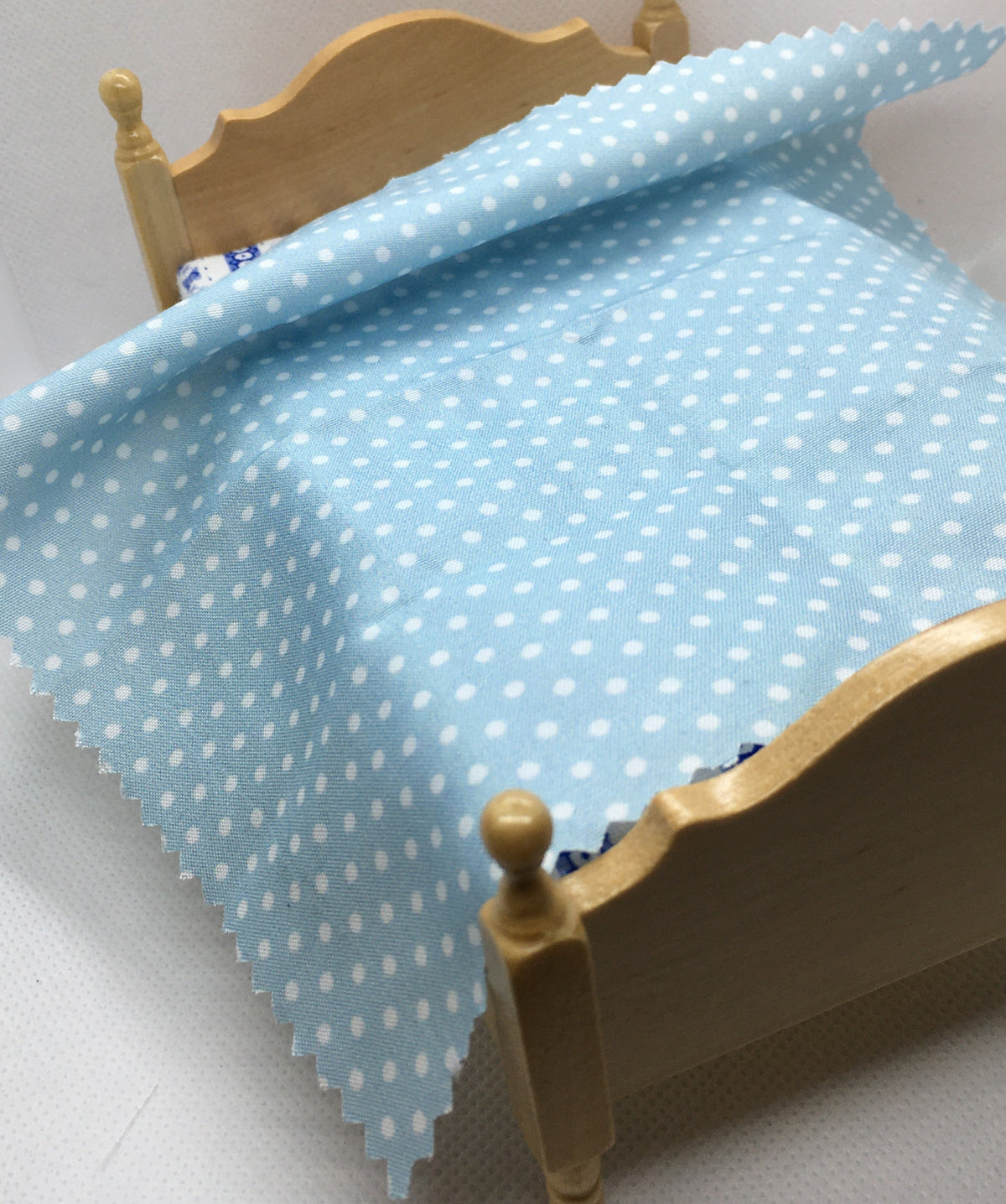 A miniature dollhouse bed with a light wood frame, styled with a light blue fabric sheet featuring a white polka dot pattern.