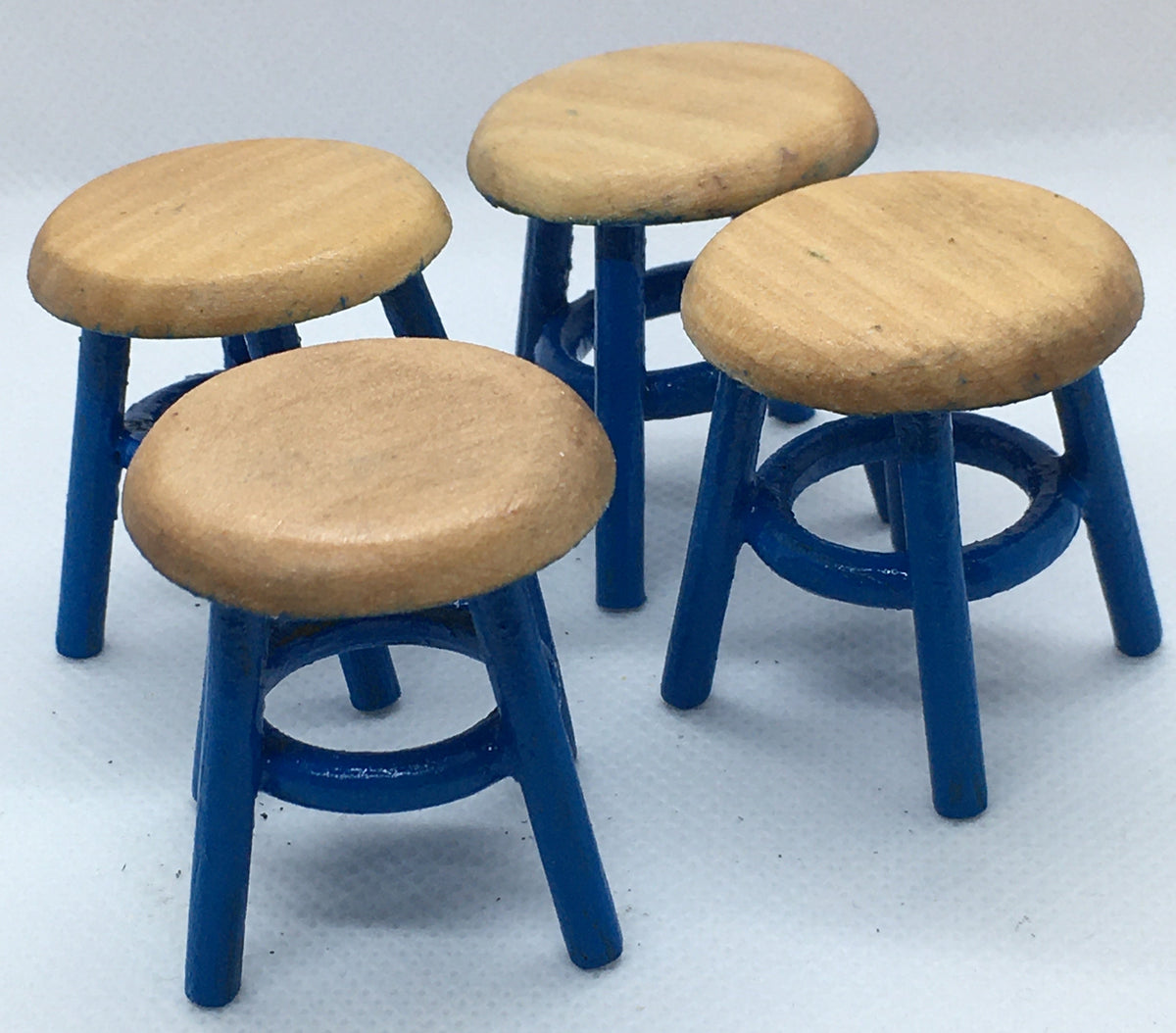 Four miniature stools with round light wood seats and blue legs, arranged on a light gray textured background.