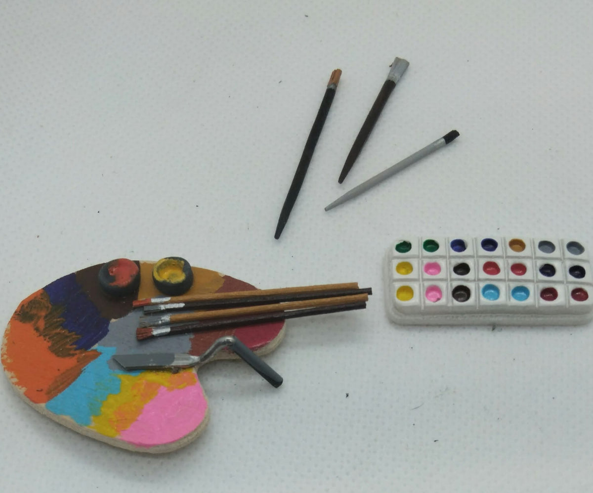 A miniature artist's set featuring a small hand-painted wooden palette with colorful paint wells, several miniature paintbrushes, a small metal mixing tool, and a miniature watercolor paint palette with 16 color wells, all arranged on a white textured background.