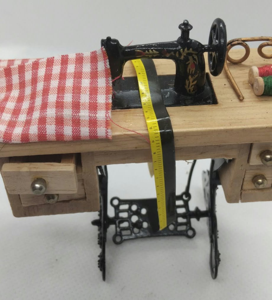 A miniature sewing machine set on a small wooden table with drawers, alongside a red-and-white checkered cloth, a yellow measuring tape, and sewing accessories, photographed against a plain light background.