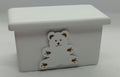 A small white rectangular box with a lid, featuring a white teddy bear charm with brown details on the front, set against a plain light gray background.