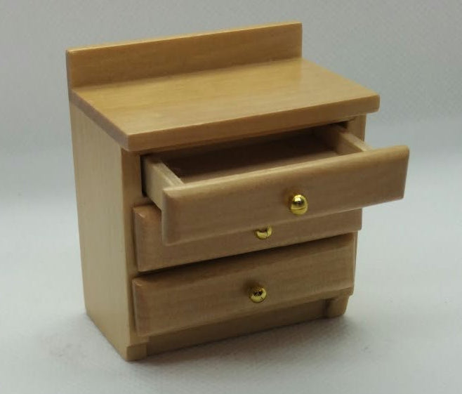 A miniature light wood bedside cabinet  with a partially open drawer and tiny gold knobs, set against a plain white background .