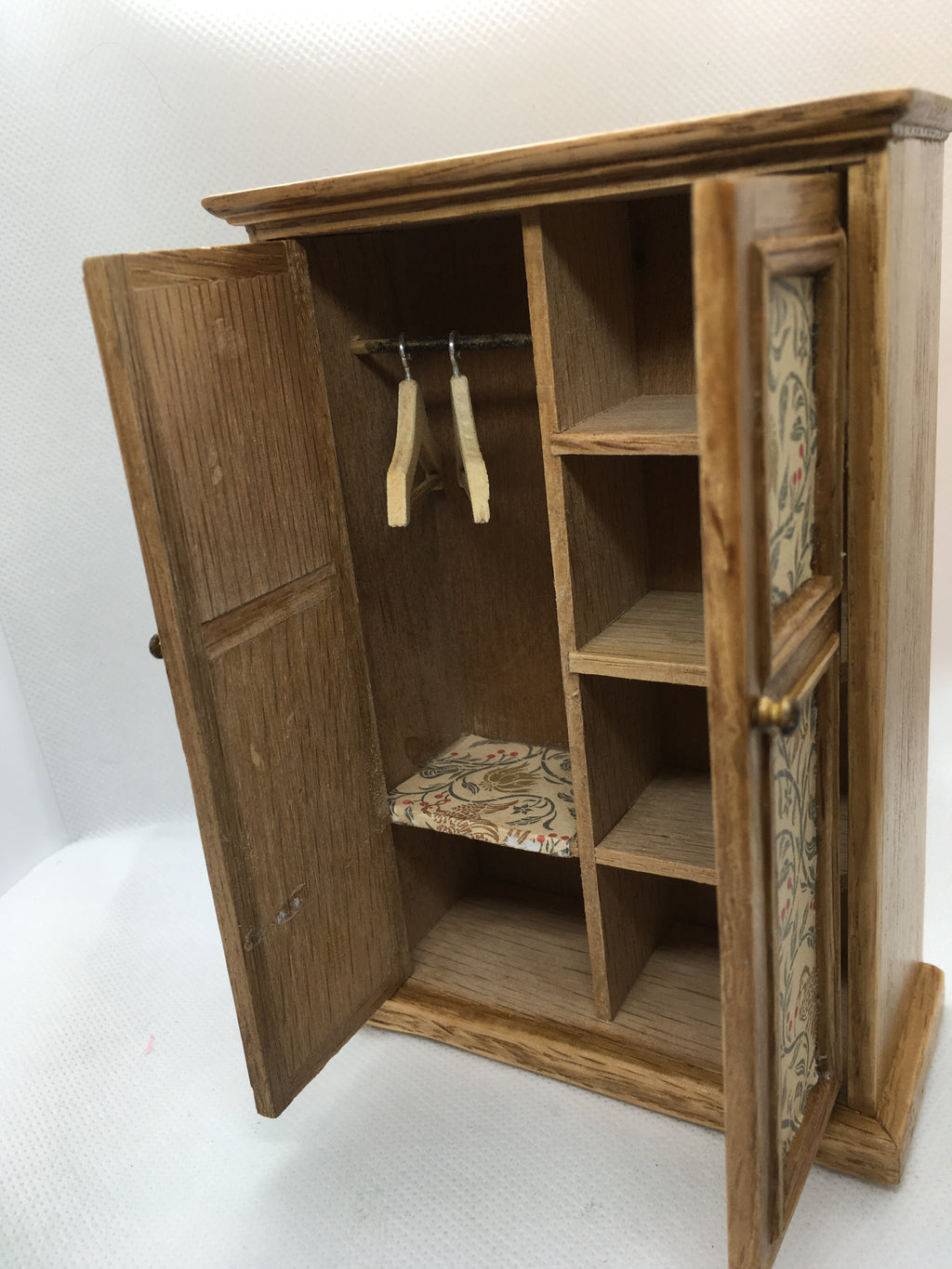 A miniature wooden dollhouse wardrobe with two doors open, showing a hanging rod with clothes hangers, interior shelves, and patterned fabric trim on the door panels.