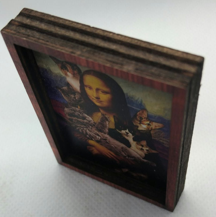 A small framed art print featuring a dark, moody painting of Leonardo da Vinci's Mona Lisa with a lot of cats, shown standing on a light gray surface.