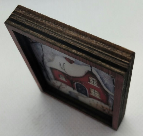A small rectangular wooden picture frame with a dark brown wood grain finish, containing a miniature red house scene, photographed on a light gray background.