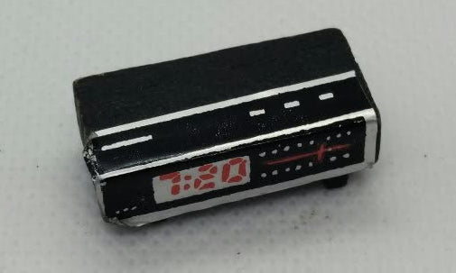 A miniature black clock radio with red and white details, photographed on a light gray textured background.
