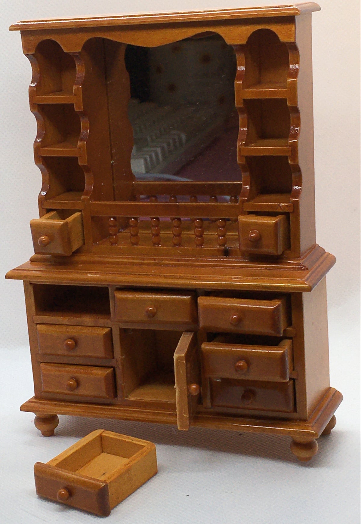 A miniature wooden vanity cabinet with an attached mirror, multiple drawers, open shelving, and one drawer pulled out in front, photographed against a plain light background.