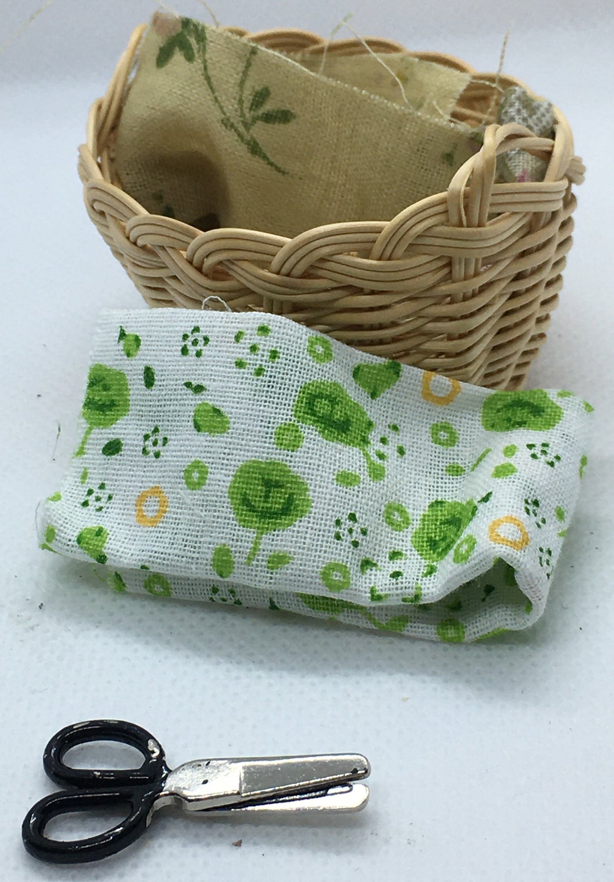 A miniature wicker basket containing beige fabric  with a green leaf pattern, placed on a white surface next to a folded white fabric piece with green and yellow floral motifs, and a pair of tiny black-handled scissors.