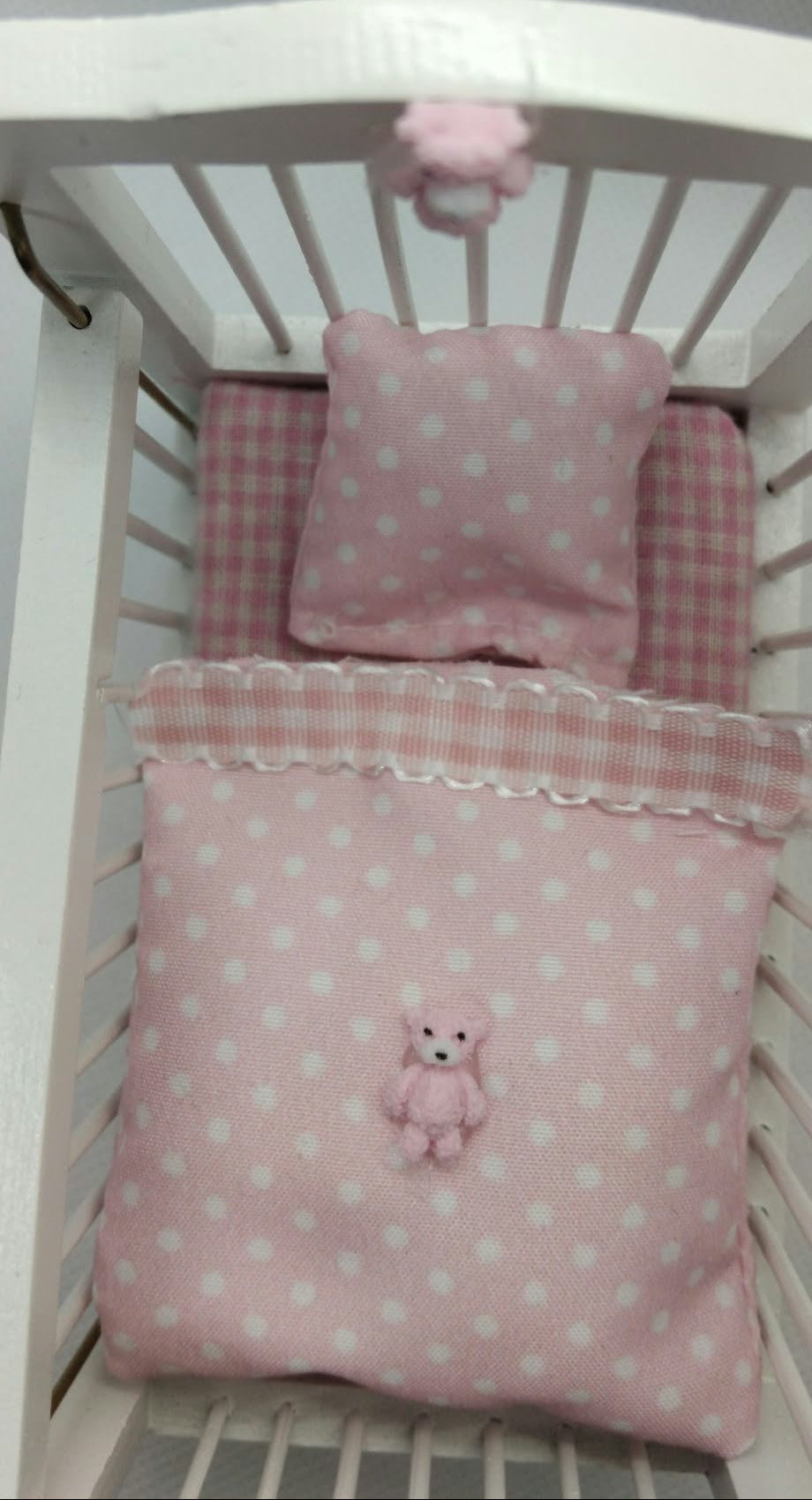 Pink baby bedding set in a white crib, featuring a pink polka dot blanket with a teddy bear applique, a matching pink polka dot pillow, and a pink and white gingham check sheet, all in miniature .