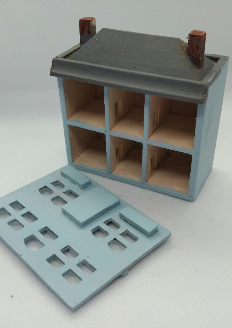 A miniature toy house model with a light blue body, dark gray roof, and brown wooden-looking details, shown with a matching detached roof panel on a plain light background.