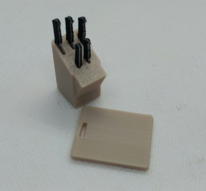 A miniature beige plastic knife block with four black knife handles inserted, shown next to a matching beige plastic chopping board on a light gray textured background.