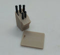A miniature beige plastic knife block with four black knife handles inserted, shown next to a matching beige plastic chopping board on a light gray textured background.