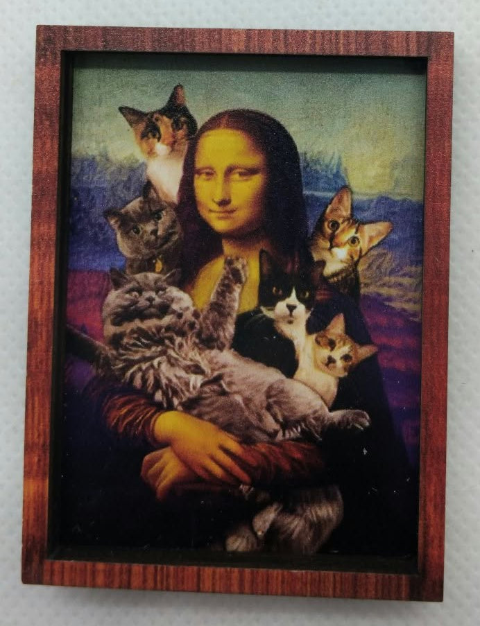 A framed art print featuring a parody of the Mona Lisa with several cats around her, displayed on a textured white wall.