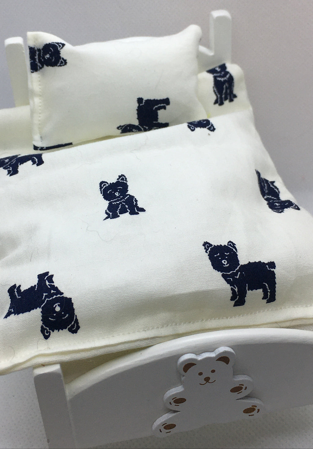 A miniature bed set with a cream-colored fabric cover and matching pillow, both printed with dark navy blue dog silhouettes, displayed on a small white wooden bed frame.