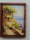 A small framed landscape painting depicting a coastal village scene with trees, stairs, and blue water, mounted on a light gray textured wall.