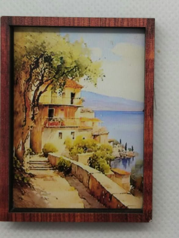 A small framed landscape painting depicting a coastal village scene with trees, stairs, and blue water, mounted on a light gray textured wall.