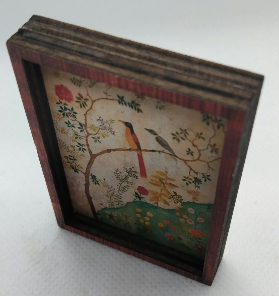 A small framed decorative artwork showing a bird perched on a flowering branch, with a dark reddish-brown wooden frame, photographed on a light neutral background.