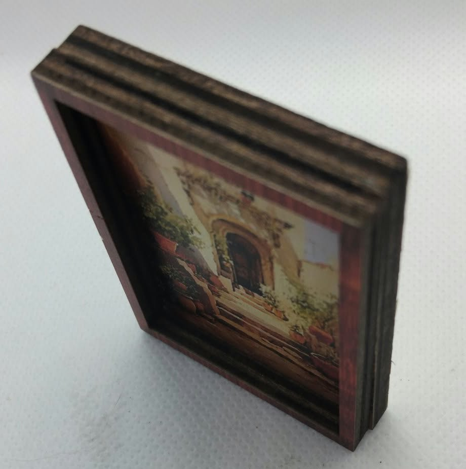 A miniature wooden frame holding a small painted scene of a building facade, photographed on a light gray textured background.