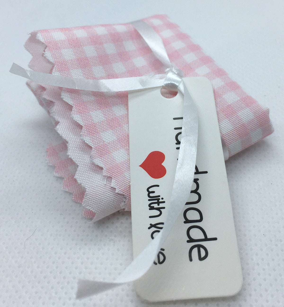 pink and white gingham check bed sheet. with a white ribbon and a white paper tag that reads ' handmade' with love and a small red heart, on a white textured background.