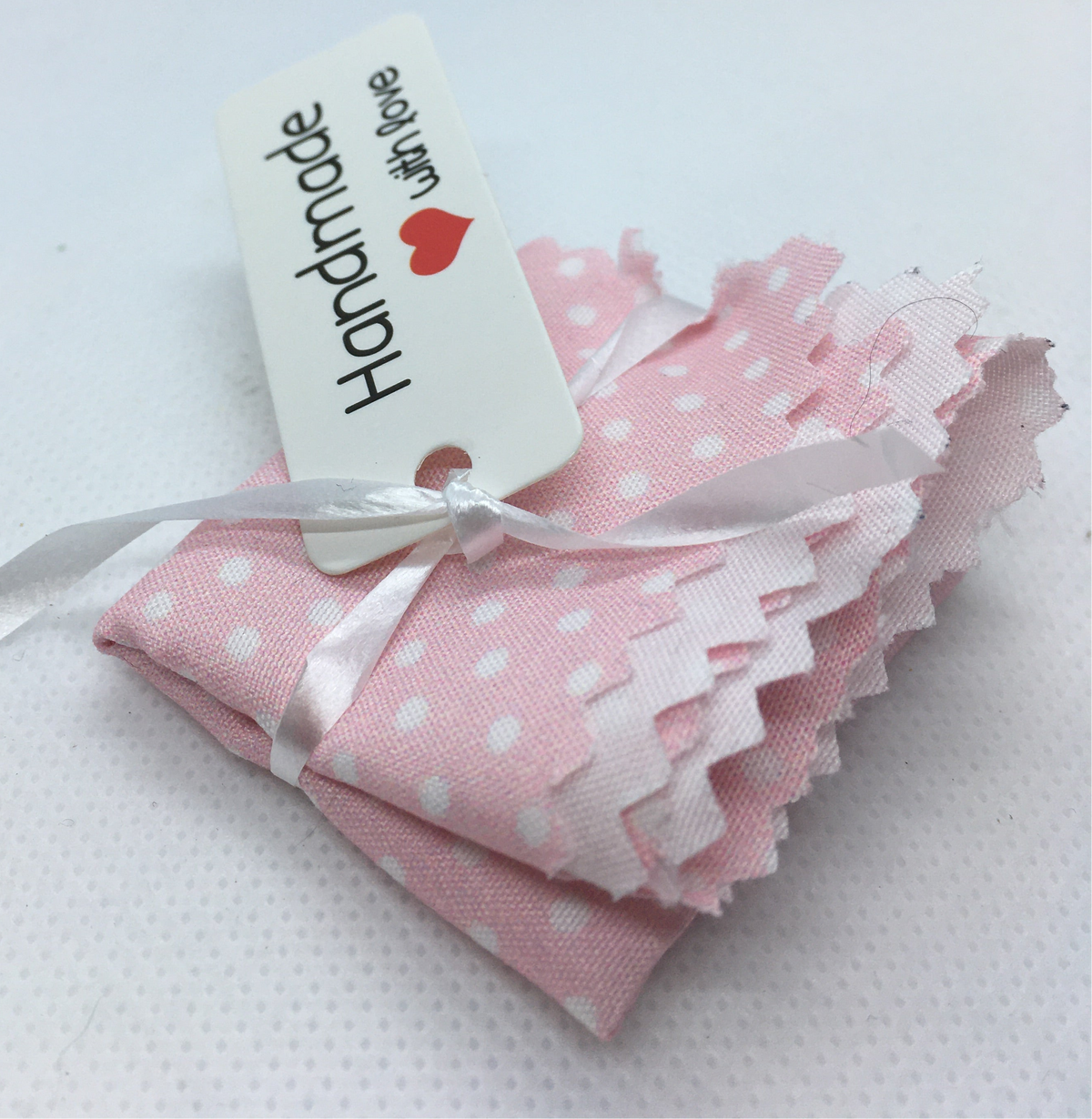 A small single bed  pink fabric bed sheet with white polka dots and ruffled edges, tied with a white ribbon and attached to a white paper tag that reads 'Handmade with love,  with a small red heart, on a white background.