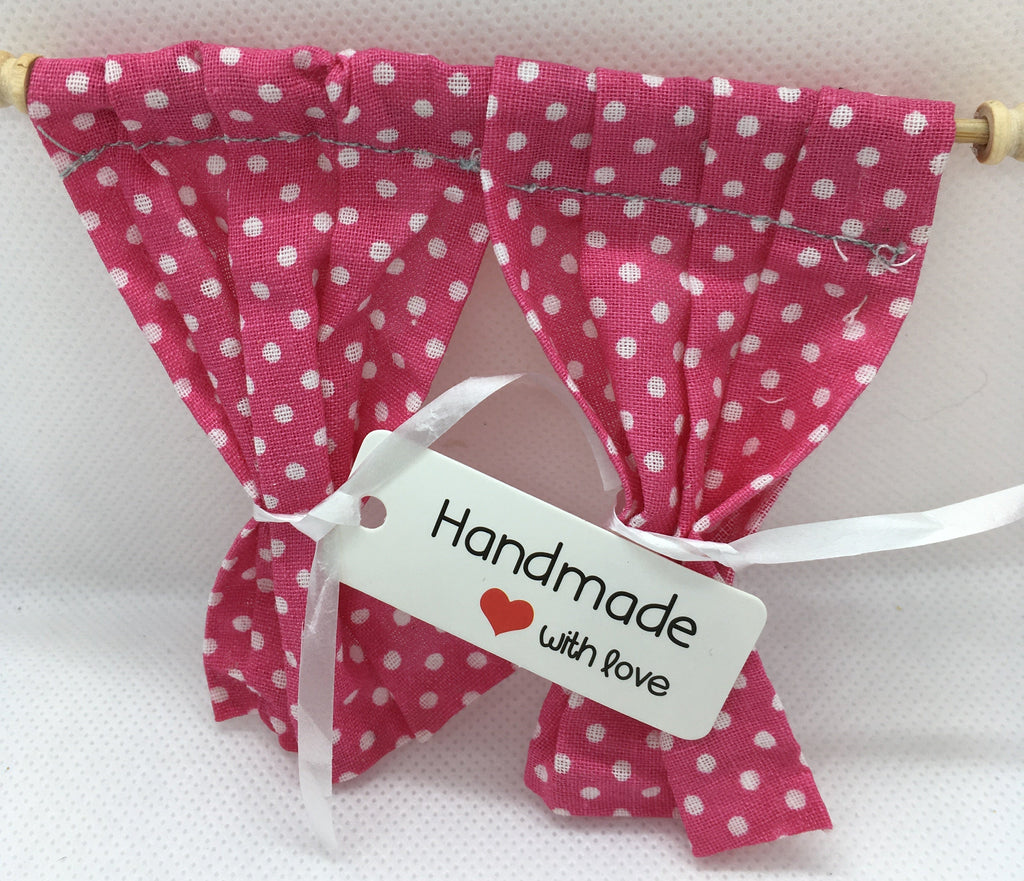 A pair of small pink fabric curtains with white polka dots hanging from a wooden rod, tied with a white ribbon and a white tag that reads 'Handmade with love'.