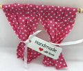 A pair of small pink fabric curtains with white polka dots hanging from a wooden rod, tied with a white ribbon and a white tag that reads 'Handmade with love'.