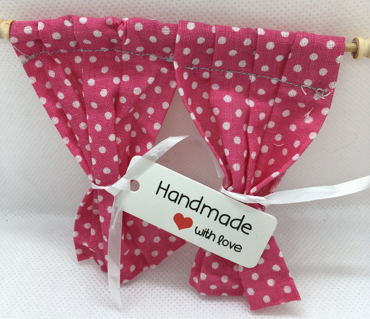 A pair of small pink fabric curtains with white polka dots hanging from a wooden rod, tied with a white ribbon and a white tag that reads 'Handmade with love'.