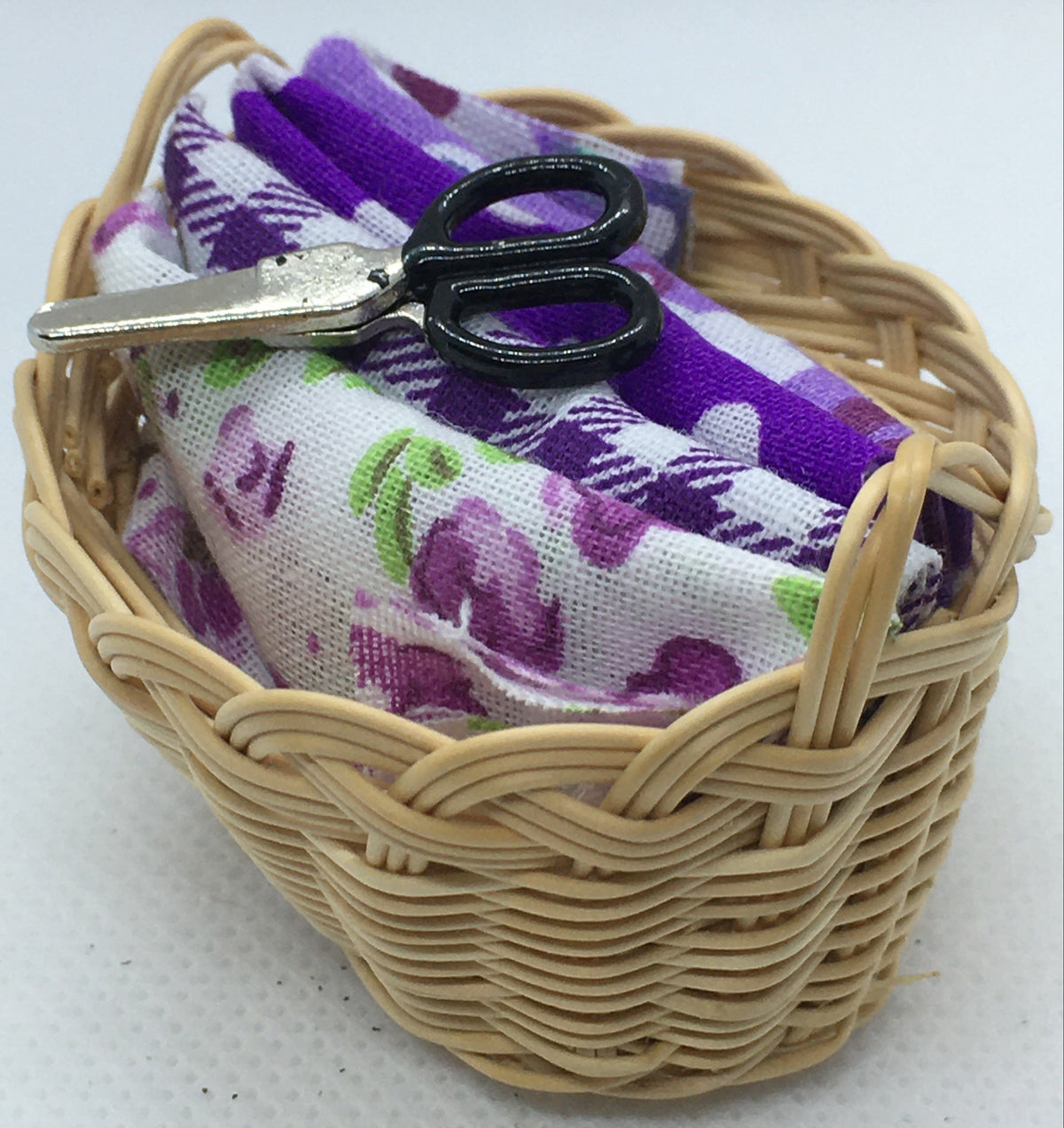 A miniature woven wicker basket holding a folded fabric with a purple and white floral pattern and a small black-handled scissors, set against a plain white background.