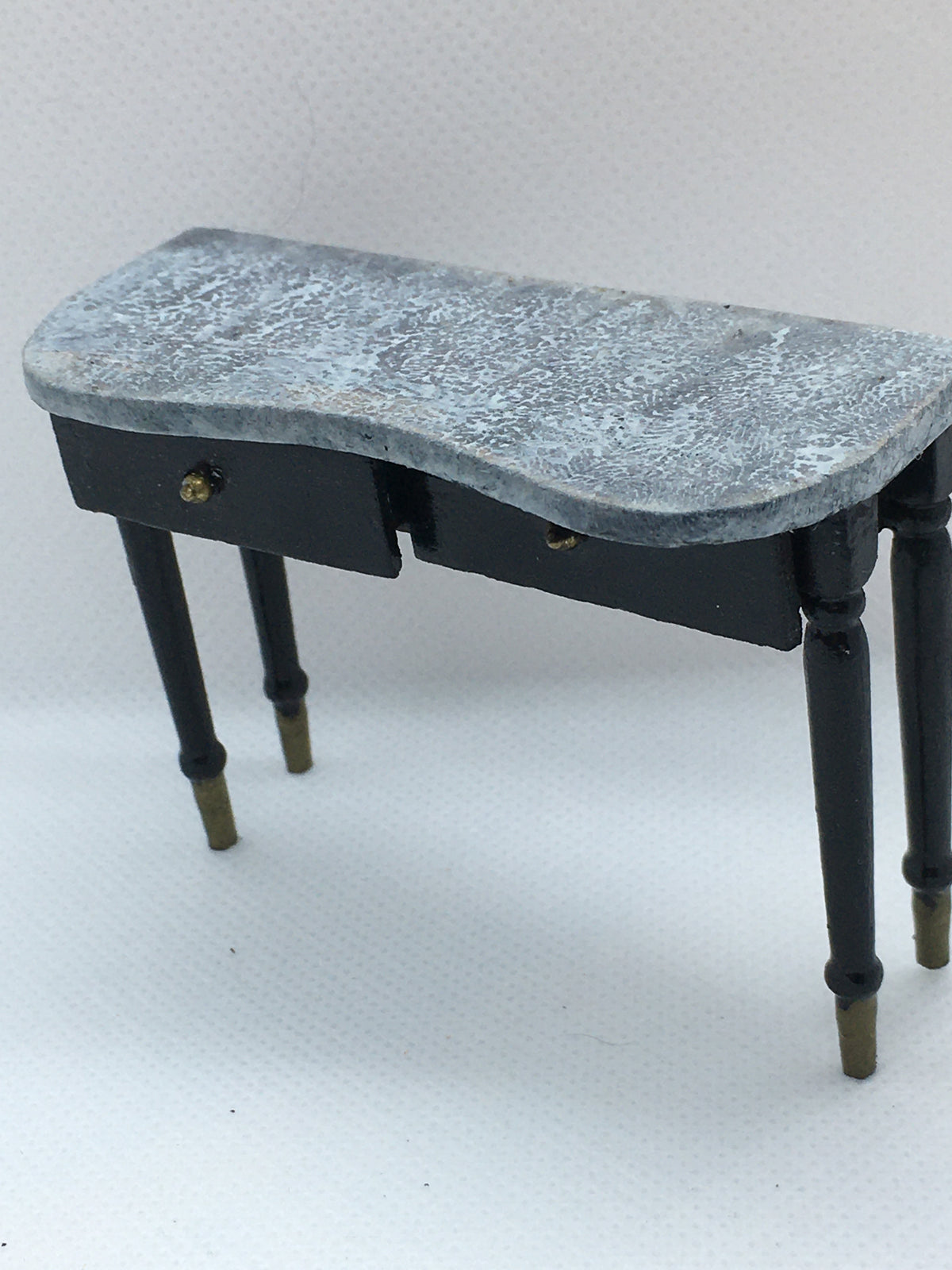 A miniature black side table with a distressed gray stone-look top, featuring two drawers and turned legs with brass-colored feet, shown on a white textured background.