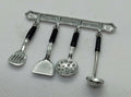 A miniature silver-colored metal kitchen utensil rack with four hanging tools, including a spatula, a scoop, a golf ball, and a ladle, each with black handles, shown on a light gray textured background.