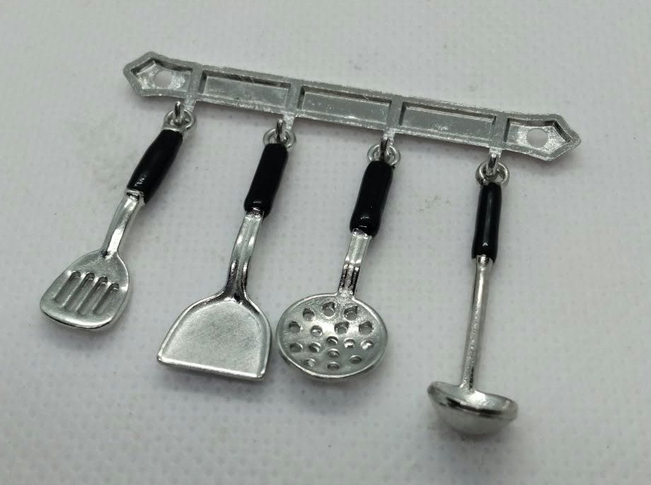 A miniature silver-colored metal kitchen utensil rack with four hanging tools, including a spatula, a scoop, a golf ball, and a ladle, each with black handles, shown on a light gray textured background.