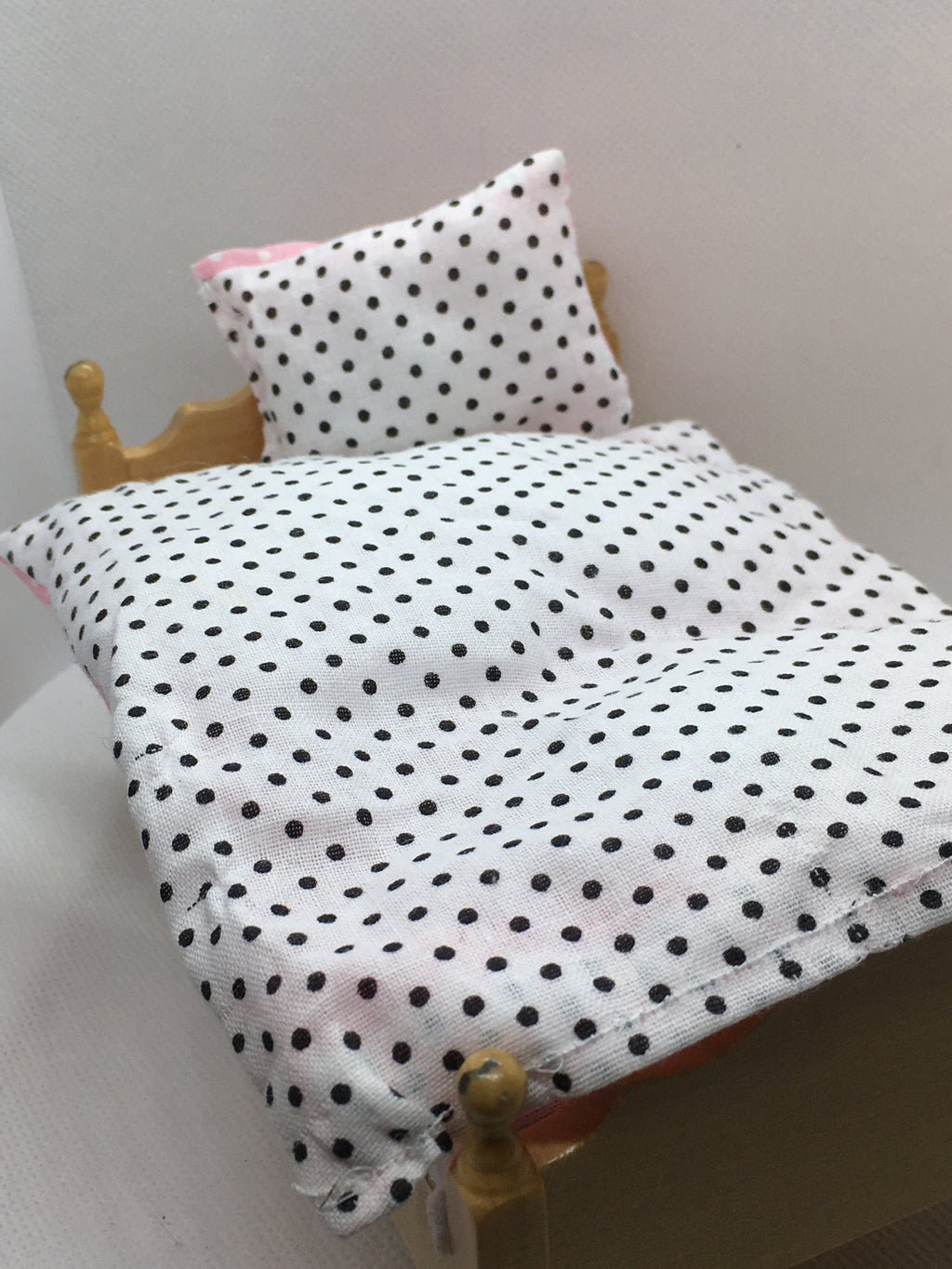 A close-up of a dollhouse bed setup featuring a white fabric duvet and matching pillow with black polka dots, resting on a miniature wooden bed frame.