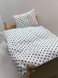 A close-up of a dollhouse bed setup featuring a white fabric duvet and matching pillow with black polka dots, resting on a miniature wooden bed frame.