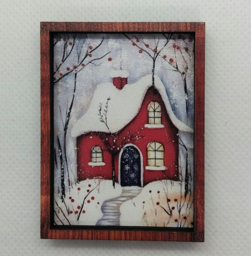 A small framed winter scene print hanging on a light gray textured wall. The artwork shows a red house with snow-covered roof and branches, a pale sky, and a snowy ground.
